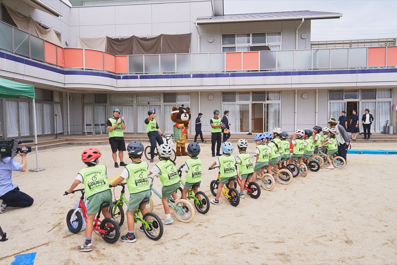 コース全体を引きで捉えた1枚。ヘルメットとビブスを着けた園児がペダルなし自転車を押して一列に並び、スタッフとくまるが見守っている