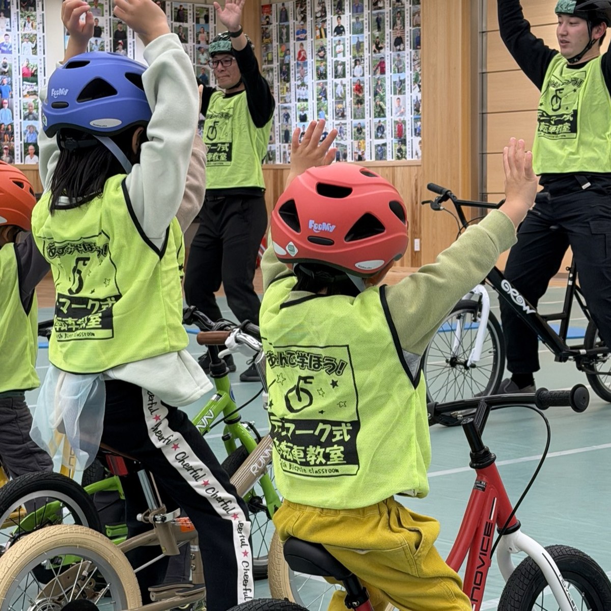 体育館のコースで、ビブスとヘルメットを着けた園児たちが自転車にまたがって両手を挙げている